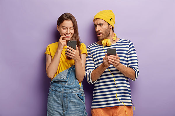 millenial-girl-looks-positively-smartphone-device-shocked-puzzled-guy-with-cellular-stand-closely-each-other-against-purple-wall-youth-with-modern-technologies-addicted-couple